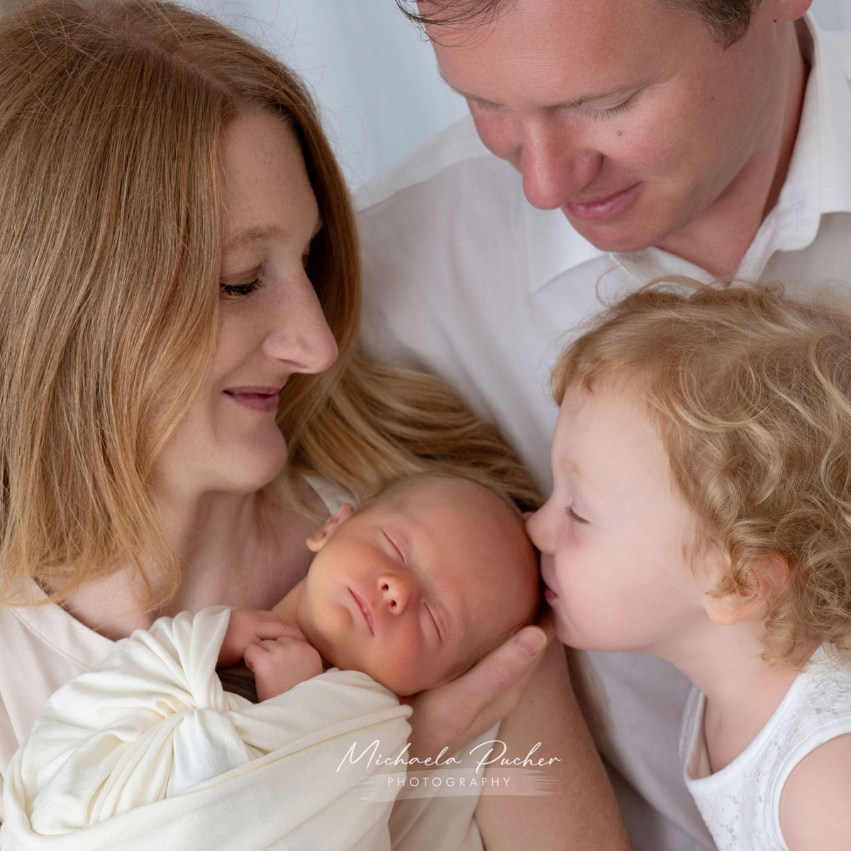 Mutter hält ihr Neugeborenes im Arm, während das ältere Geschwisterkind dem Baby liebevoll einen Kuss gibt und der Vater lächelnd auf seine Familie blickt.